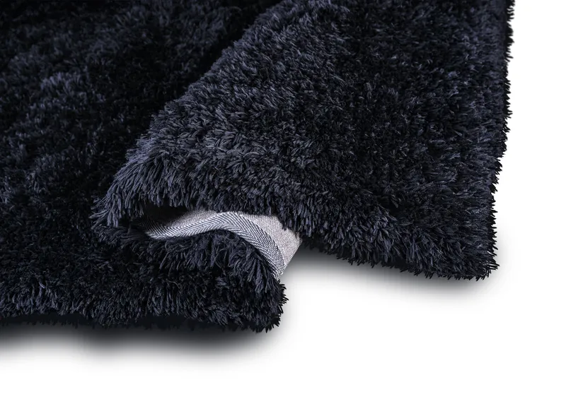 Woven Bliss – A plush, shaggy black rug, revealing its thick, soft texture. The rug is slightly folded, exposing a bit of its underside, which is a light grey woven material