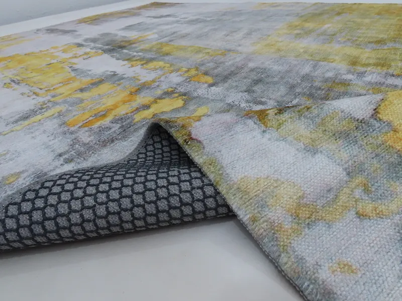 Woven Bliss – A rug with an abstract design in grey and yellow tones. The edge of the rug is lifted, revealing a grey backing with a hexagonal pattern.