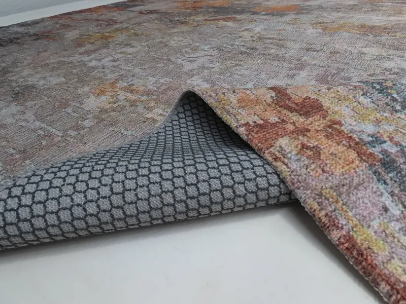 Woven Bliss – A rug with an abstract design in brown, beige, and rust tones. The edge of the rug is lifted, revealing a grey backing with a hexagonal pattern.